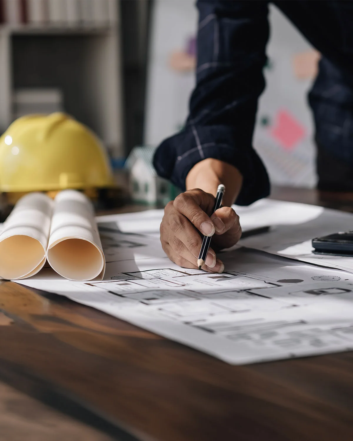 An engineer's hands on a design drawing, pencil, yellow hard hat and rolled-up plans on a wooden table