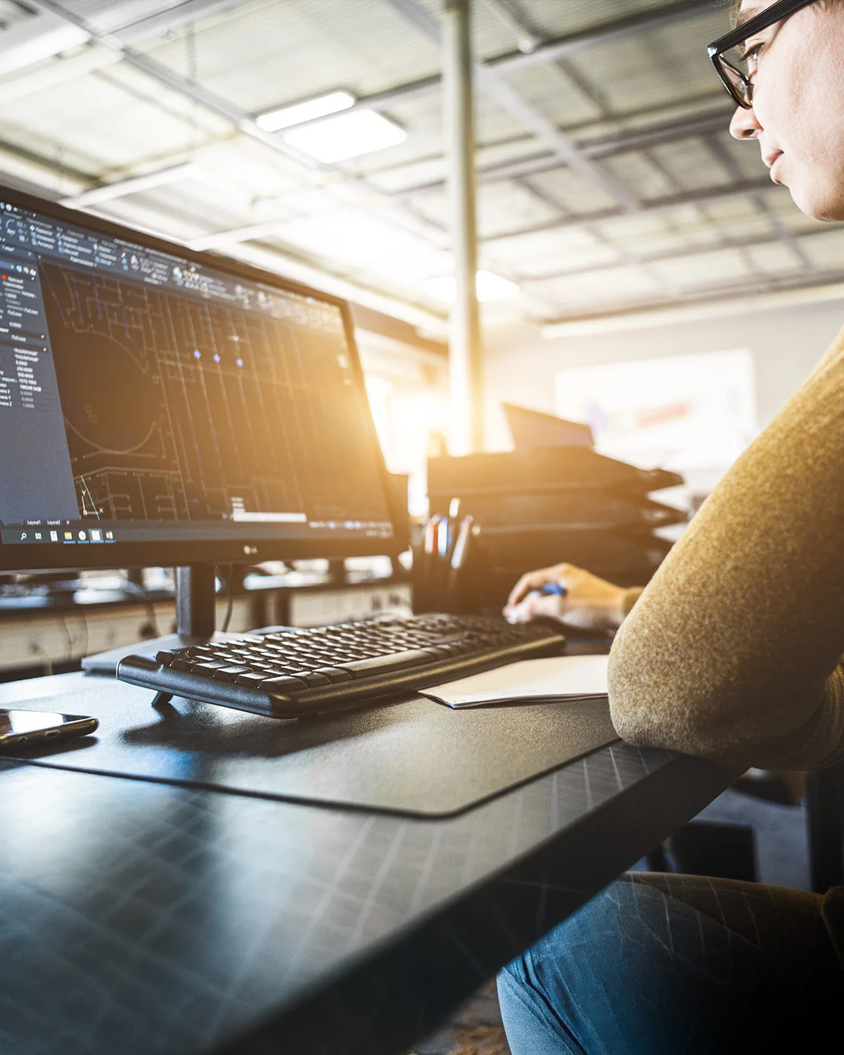 Engineer at a desk in front of a large monitor showing an AutoCAD drawing, industrial hall in the background